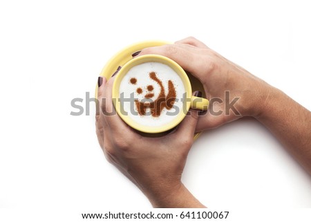 Hand holding a cute yellow cup with coffee cream. Food art creative concept image, Panda Bear shape drawing with cinnamon powder over white background.