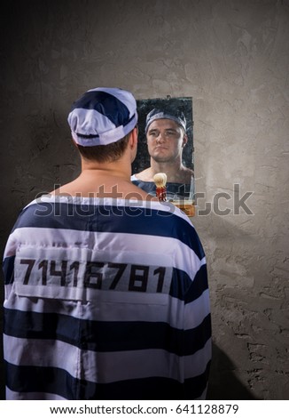 Angry prisoner wearing  prison uniform with sewed number standing and looking at his reflection in the mirror with shave brush on the shelf in a prison cell