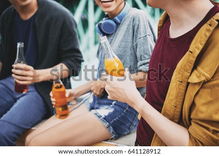 Group of cheerful friends chatting animatedly with each other while sitting on bridge railing and drinking delicious cocktails, close-up shot