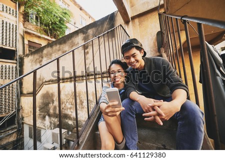 Two smiling Asian friends gathered together in urban slums and taking selfie on modern smartphone