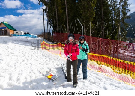 Two young women in ski suits, with helmets and ski goggles standing near the fence in a ski-resort in winter period