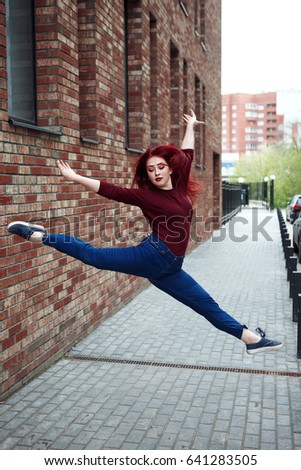 Beautiful serious Shapely young Caucasian woman dancer jumping against brick wall on the street In jeans and a burgundy jacket full length looking down outdoors