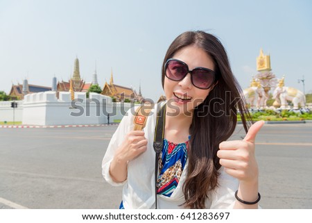 happy female backpacker visiting Bangkok awesome Grand Palace and showing thumb up gesture taking picture with building on Thailand travel.
