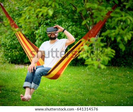 Young bearded man wearing virtual reality goggles relaxing in a garden hammock. Lifestyle VR fun and relax concept