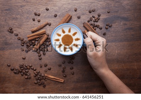 Hand holds beautiful blue mug with coffee cream. Food art creative concept image, SUN shape drawing with cinnamon powder over wooden background.