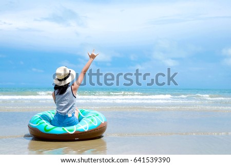 Woman sitting on swim ring with love hand sign gesture over blue sky at summer beach background, travel, holiday vacation concept