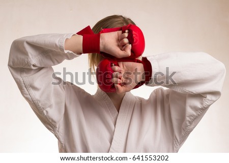 Young woman in kimono on white background, blonde hair, karate, covers her face with her hands, hands in red gloves