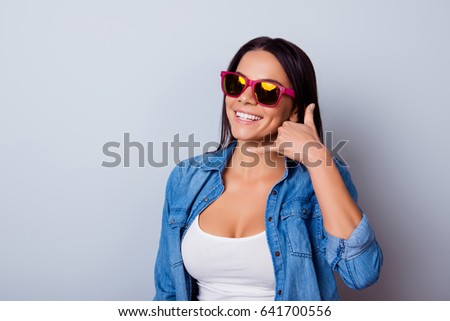Cheerful latin girl with beaming smile is gesturing to call her with a hand. She is wearing casual jeans shirt and white singlet, sunglasses and stands on a light background