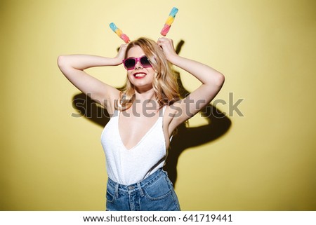 Picture of young lady with bright lips makeup standing over yellow background with ice cream wearing sunglasses. Looking at camera.
