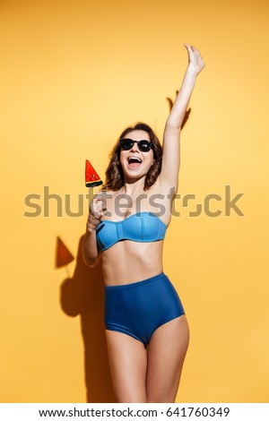 Image of cheerful young lady in swimwear isolated over yellow background. Looking aside eating candy.