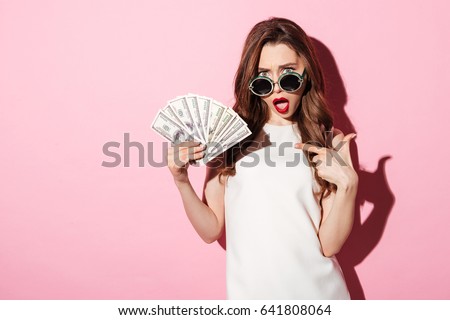 Photo of a confused young brunette woman in white summer dress holding money pointing and looking at camera over pink background.