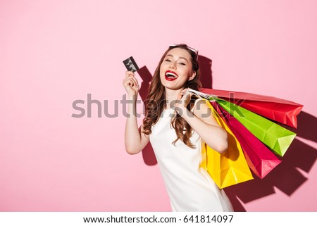 Picture of a cheerful young brunette woman in white summer dress holding credit card posing with shopping bags and looking at camera over pink background.