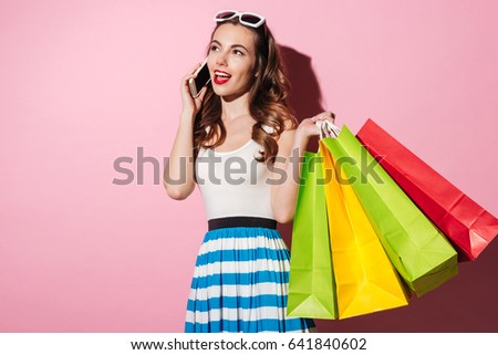 Portrait of a young smiling woman holding colorful shopping bags and talking on mobile phone isolated over pink background