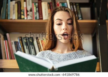 Picture of young concentrated woman student sitting in library reading book. Looking aside.