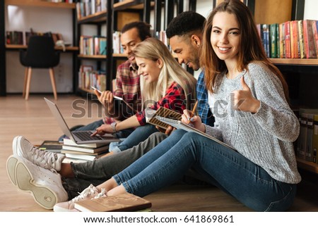 Picture of young students sitting in library on floor using laptop computer and reading book. Woman looking at camera showing thumbs up.