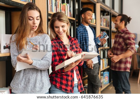 Photo of young smiling women students standing in library reading books. Looking aside.