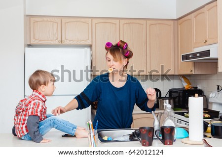 Busy white Caucasian young woman mother housewife with hair-curlers in her hair cooking preparing dinner meal in kitchen, child son boy sitting on table, crazy parent life Royalty-Free Stock Photo #642012124