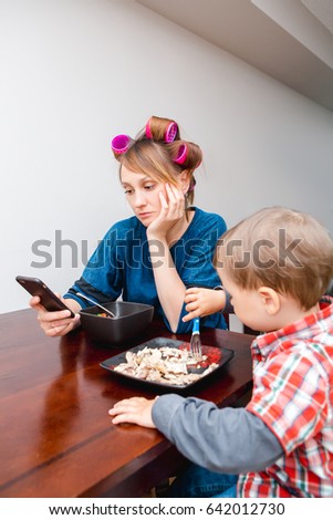 Bored tired white Caucasian young woman mother housewife with hair-curlers in hair looking on phone surfing Internet, f child son boy sitting eating meal lunch, lifestyle concept