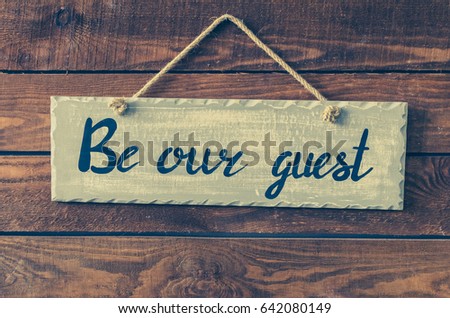 Be our guest - wording on board hanging on a rustic wooden background. Welcome concept. Toned image.