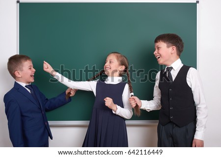 child play and having fun, boys pull the girl braids, near blank school chalkboard background, dressed in classic black suit, group pupil, education concept