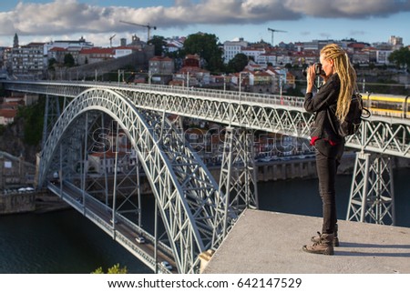 Traveller girl photographer with blonde dreadlocks is takes photo Dom Luis I bridge over Douro river, Porto, Portugal.