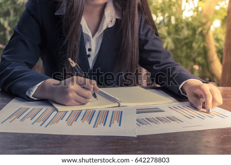 Close up of young business woman wearing black suit working and writing in notebook at coffee shop, business summary charts on wooden table. Golden time with beautiful light background.