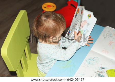 A small child looks at the pictures in the children's book at the children's table. A girl is reading a children's book in the children's room.