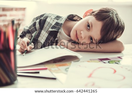 Handsome schoolboy is looking at camera and leaning on the table while drawing in his room at home