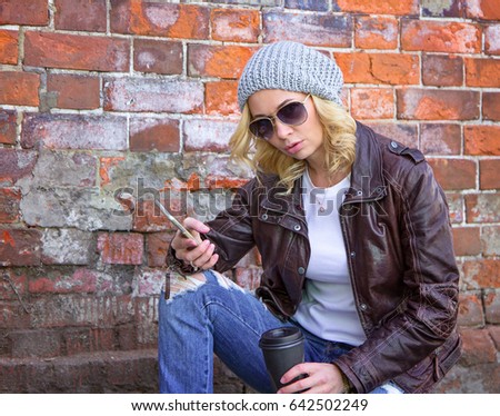 Young stylish pretty woman wearing jeans, leather jacket and sunglasses using smart phone app with coffee to go with brick wall background. Urban lifestyle