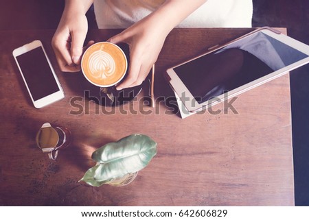 Top view of woman hands holding hot latte coffee with tablet and smartphone on wooden table in the cafe