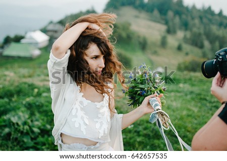 Beautiful bride in white dress on a green field in the mountains, on grass. bouquet and wreath of wild flowers. Wind driveth brown hair. A photographer takes pictures of bride. Hipster. Rustic. Happy.