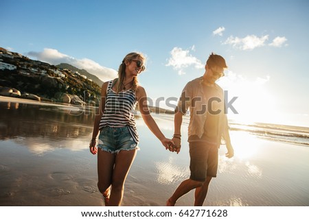 Outdoor shot of loving young couple walking on the sea shore holding hands. Young man and woman walking on the beach together.