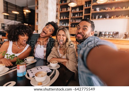Young man taking selfie with friends making funny faces in a cafe. Crazy young people in a coffee shop making a self portrait.