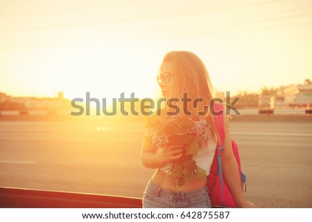 Young attractive hipster girl stands at the highway and holds a bouquet of lilac flowers. She is dressed in top and jeans. Backlight photography.
