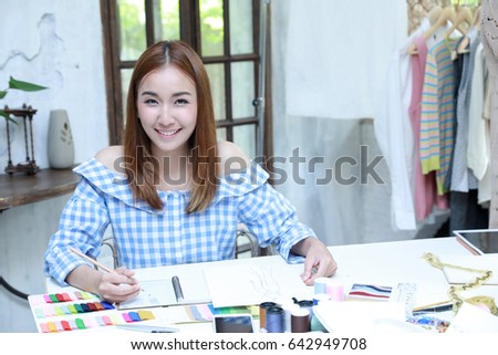 Cheerful asian designer working on her beautiful dress at costume design desk in factory studio .Asian fashion woman designer drawing design sketch working in her manufacturing office studio.