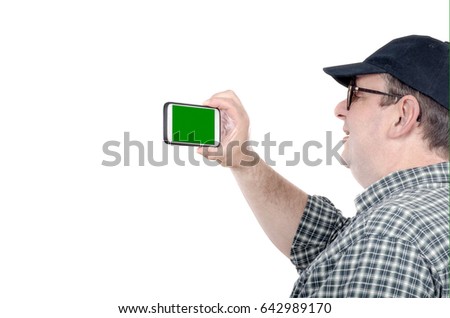 Fat man with chubby face shoots with mobile phone. Fifty years old man looks at chroma key screen. He wears plaid shirt and black cap. Tourism concept. Horizontal mid shot on white background