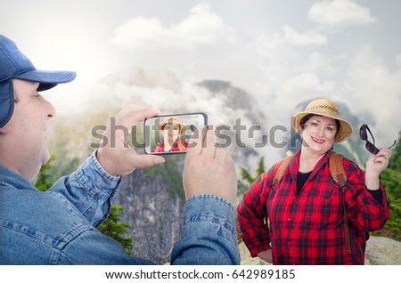Retired couple tourists backpacking in mountains. Man takes pictures at his cheerful partner with cellphone. Elderly woman in red checkered shirt poses on blurred outdoors background. Travel concept
