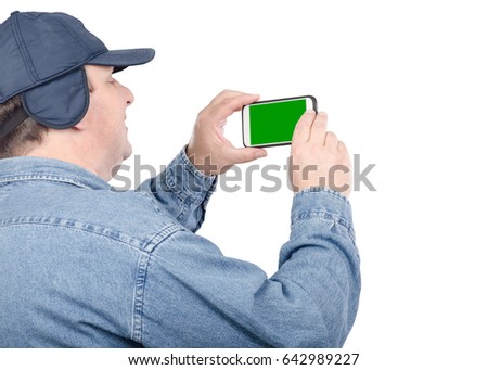 Fifty years old man shoots with cellphone. Fat man with chubby face looks at chroma key screen. He wears denim shirt and navy cap with ear flaps. Tourism concept. Mid shot on white background