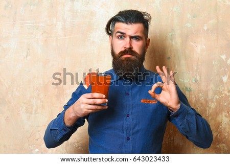Bearded man, long beard. Brutal caucasian serious hipster with moustache in denim shirt holding tropical alcoholic fresh cocktail with orange piece on texture background