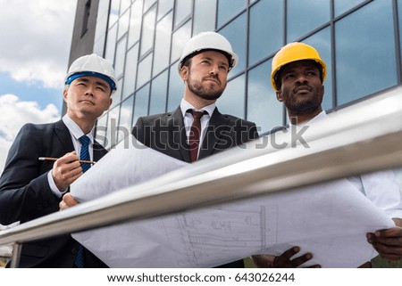 Multiethnic group of professional architects in helmets working with blueprint outside modern building