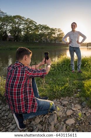 Boy taking photo of happy smiling girl in the park near the river. Summer sunny day