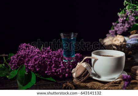 Cup of coffee on linen napkin and several macarons in braid vase with branch of lilac and pile of absinthe