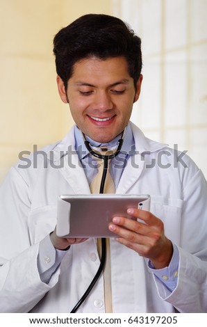 Handsome smiling doctor with an Stethoscope holding from his neck, checking something in his tablet, in office background