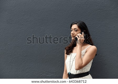 Close up of beautiful brunette female model serious face speaking on cell phone in white dress and black belt while standing against grey dark background