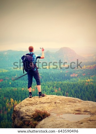 Amateur photographer prepare camera to takes impressive photos of  misty fall mountains. Tourist photographer at sharp rocky edge on high view point, colorful misty forest bellow.