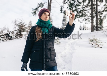 The lovely girl in a lilac cap, a blue jacket and a menthol scarf takes a selfie. She gently smiles. On the street winter.