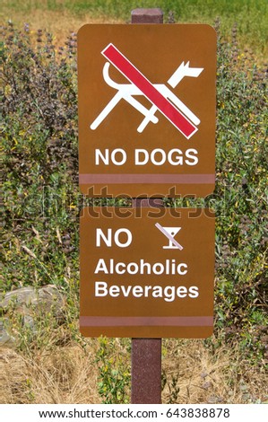 Brown signs, no dogs and no alcoholic beverages. Tall weeds growing in background.