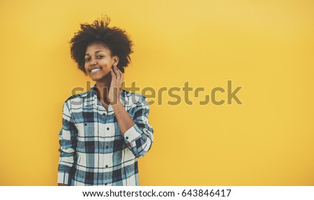 Young biracial student girl with perfect smile and curly afro hair in casual checkered outfit staying in front of solid yellow wall and touching her face, with copy space place for your advertising
