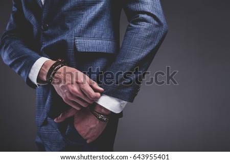 Fashion portrait of young businessman handsome model man dressed in elegant blue suit with accessories on hands posing on gray background in studio. Hands in pockets Royalty-Free Stock Photo #643955401