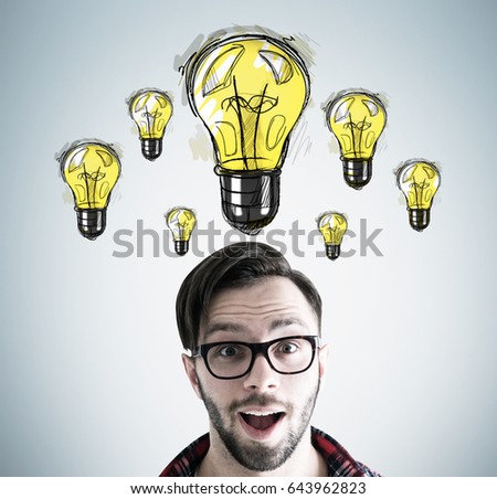 Close up of an astonished hipster guy s head with many yellow light bulbs drawn above it on a gray wall behind